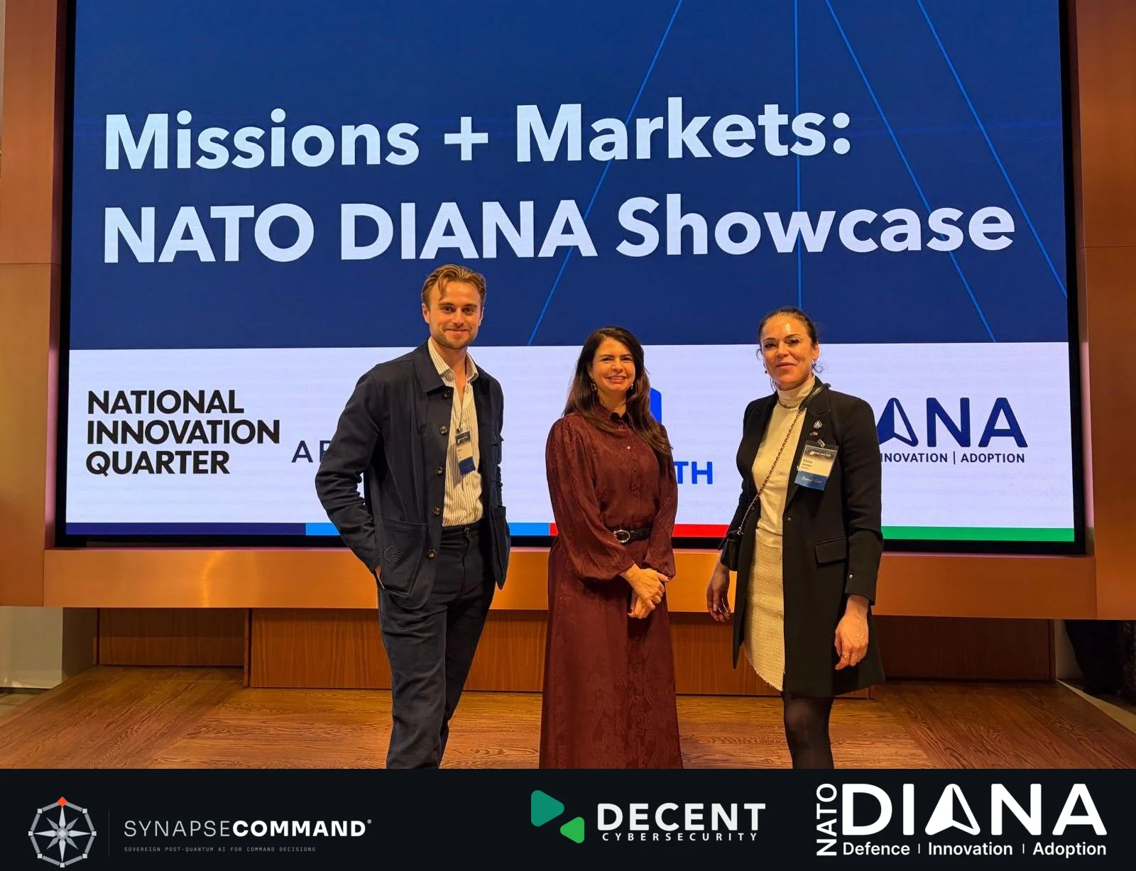 Three attendees in front of the Missions + Markets NATO DIANA Showcase display, with SynapseCommand, Decent Cybersecurity and NATO DIANA branding beneath the photograph.